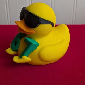 May include: A yellow rubber duck wearing black sunglasses and holding a green number 67. The duck has an orange beak and is sitting on a red surface. The duck is a fun, novelty item.