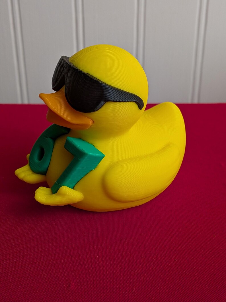 May include: A yellow rubber duck wearing black sunglasses and holding a green number 67. The duck has an orange beak and is sitting on a red surface. The duck is a fun, novelty item.