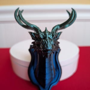 May include: A 3D-printed wall mount featuring a stylized dragon head with large antlers. The sculpture is primarily black with teal highlights, mounted on a shield-shaped plaque. The piece is displayed on a white surface against a red background.