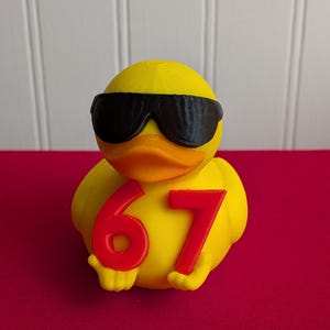 May include: A yellow duck figurine wearing black sunglasses and holding the number 67 in red. The duck has an orange beak and is set against a red background.