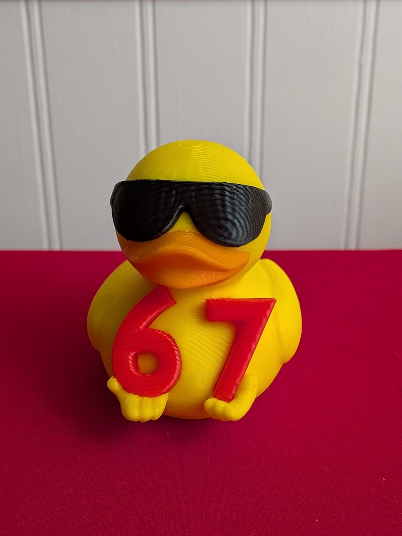 May include: A yellow duck figurine wearing black sunglasses and holding the number 67 in red. The duck has an orange beak and is set against a red background.