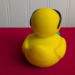 May include: A bright yellow rubber duck with black headphones. The duck is sitting on a red surface. The duck is a solid color and has a smooth texture.
