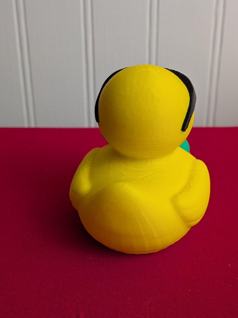 May include: A bright yellow rubber duck with black headphones. The duck is sitting on a red surface. The duck is a solid color and has a smooth texture.