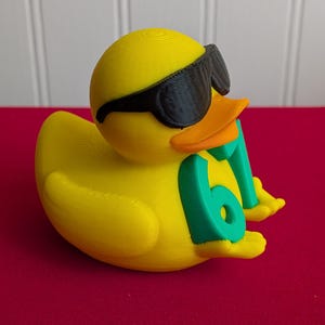 May include: A yellow rubber duck wearing black sunglasses and holding a green number 61. The duck has an orange beak and is resting on a red surface. The duck is a fun, novelty item.