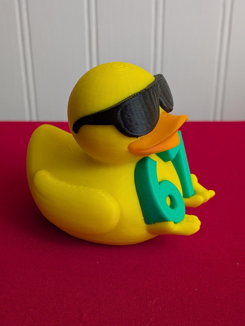 May include: A yellow rubber duck wearing black sunglasses and holding a green number 61. The duck has an orange beak and is resting on a red surface. The duck is a fun, novelty item.