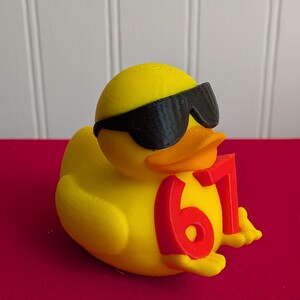 May include: A yellow rubber duck wearing black sunglasses and holding the number 67 in red. The duck has an orange beak and is sitting on a red surface. The background is white.