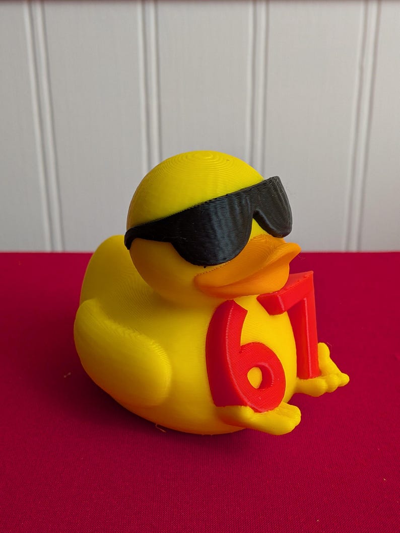 May include: A yellow rubber duck wearing black sunglasses and holding the number 67 in red. The duck has an orange beak and is sitting on a red surface. The background is white.