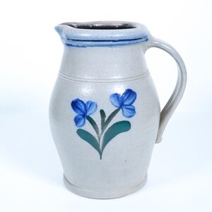 May include: A light gray stoneware pitcher featuring a hand-painted blue floral design with green leaves on the front. The vessel has a curved handle, a wide rim with a pouring spout, and a smooth, glazed finish throughout the exterior surface.