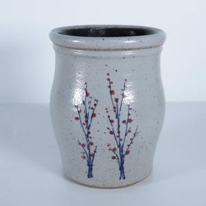 May include: A speckled, off-white ceramic utensil holder with a dark rim and interior. Two stylized blue and red floral designs are painted on the front. The holder has a slightly curved shape.