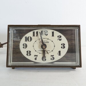 May include: A vintage rectangular Sunbeam Light Dial clock with a brown frame and a clear plastic cover. The clock face is oval, cream-colored, with black numerals and hands. The background has a wood grain pattern.