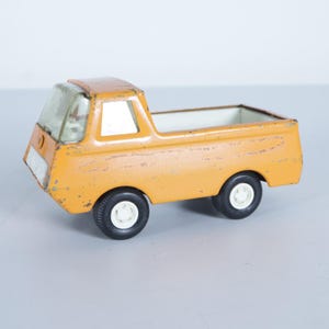 May include: Vintage orange toy pickup truck with black tires and white rims. The truck has a clear windshield and a white interior bed. The toy truck is made of metal and shows signs of wear.
