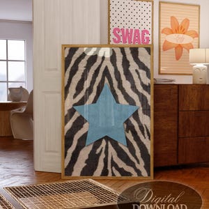 May include: A rectangular wall art print in a wooden frame featuring a black and white zebra stripe pattern with a large, centred, solid light blue five-pointed star. The print is displayed in a room setting with other framed art pieces.