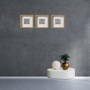 Puede incluir: Tres impresiones cuadradas enmarcadas con caligrafía árabe en un interior moderno. Las impresiones están en marcos de madera clara con paspartús blancos. Una esfera blanca y una pequeña planta en maceta en una maceta dorada se encuentran sobre una mesa ovalada blanca.