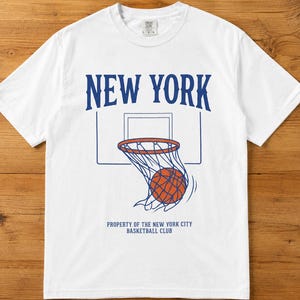 Op de afbeelding: Wit T-shirt met een basketbalafbeelding. Het ontwerp toont de tekst "NEW YORK" boven een basketbalnet met een bal. Daaronder staat de tekst "PROPERTY OF THE NEW YORK CITY BASKETBALL CLUB."