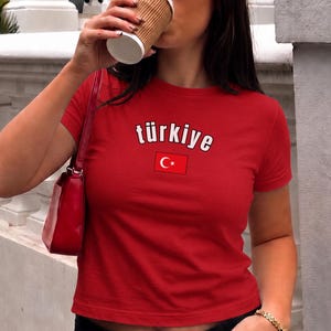 Peut inclure: Haut court rouge avec le mot "türkiye" en blanc et un petit drapeau turc. Le t-shirt à manches courtes est porté avec un pantalon noir. La personne porte des lunettes de soleil et tient une tasse de café.