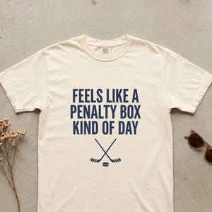 May include: Ivory-colored t-shirt with the text "FEELS LIKE A PENALTY BOX KIND OF DAY" in navy blue. Below the text are crossed hockey sticks and a puck. Sunglasses are on the right side of the shirt.