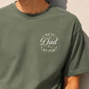 Könnte beinhalten: Ein olivgrünes Kurzarm-T-Shirt mit Rundhalsausschnitt und einem weißen Aufdruck auf der linken Brust. Die Grafik zeigt eine Golffahne und den Text "BEST DAD BY PAR" in einem kreisförmigen Design mit dekorativen Blättern um den Schriftzug.
