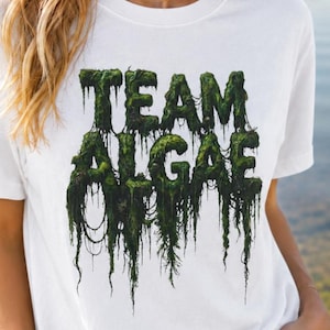 Peut inclure: Un t-shirt blanc à manches courtes avec le texte "TEAM ALGAE" imprimé dans une police texturée vert foncé, rappelant la mousse, qui semble dégouliner de végétation aquatique suspendue. Le motif est centré sur la poitrine du vêtement.