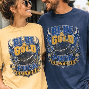 Op de afbeelding: Een stel draagt bijpassende t-shirts met een hockeythema. De shirts zijn geel en marineblauw en hebben een hockeypuck en de woorden "Blue & Gold Puck Culture" in een vintage stijl. Ze lachen en dragen zonnebrillen.