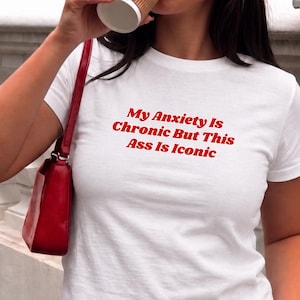 Puede incluir: Top corto blanco con el texto rojo "My Anxiety Is Chronic But This Ass Is Iconic." La modelo lleva vaqueros negros y gafas de sol, sosteniendo una taza de café y un bolso rojo. La palabra "white" está en la esquina inferior derecha.