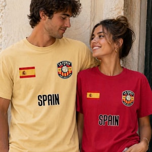 May include: Two people wearing matching t-shirts. One shirt is yellow, the other is red. Both shirts have the Spanish flag, a soccer emblem, and the word "SPAIN" printed in black. The yellow shirt is paired with light-colored pants, and the red shirt is paired with white shorts.