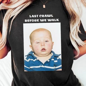 Può includere: T-shirt nero con la scritta "LAST CRAWL BEFORE WE WALK" sopra una foto di un bambino con una camicia a righe blu e bianche. Il bambino ha un occhio chiuso.