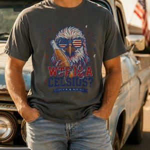 May include: A charcoal gray t-shirt featuring a graphic of a bald eagle wearing American flag sunglasses and holding a glass bottle. The text reads "WTF IS A CELSIUS?" in red and blue lettering, accented by small lightning bolts and firework designs.