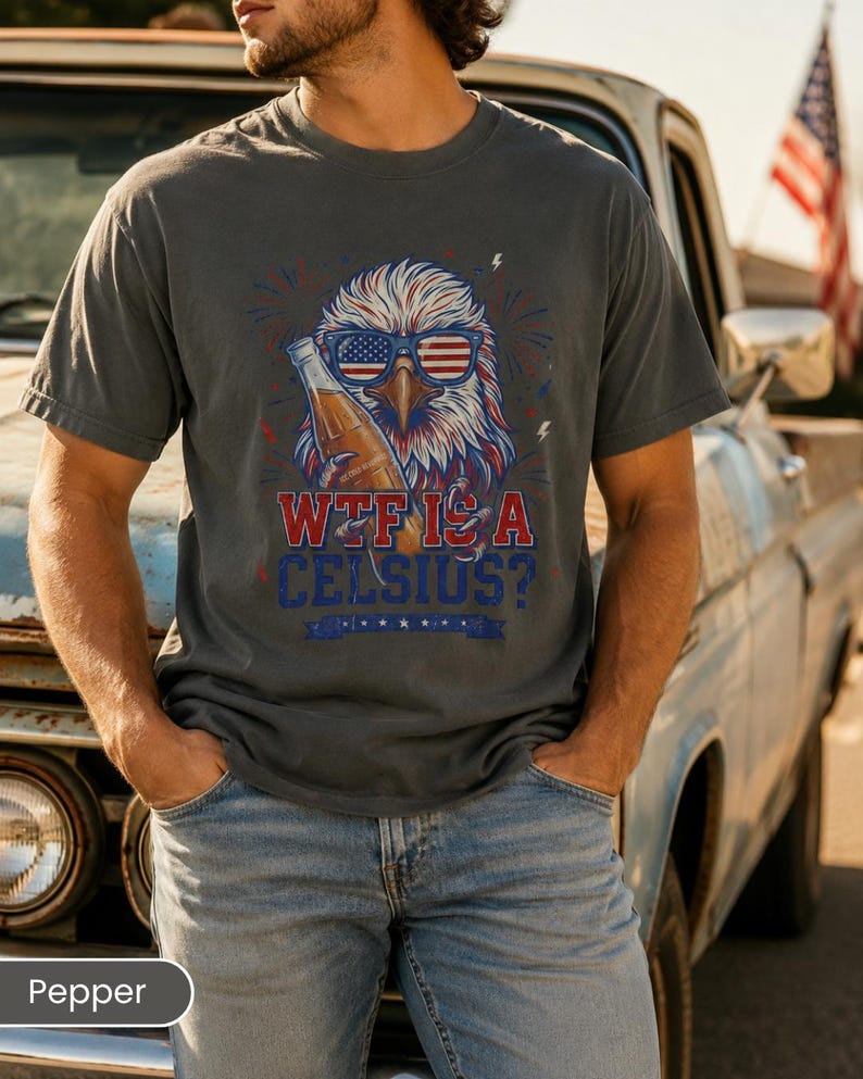 May include: A charcoal gray t-shirt featuring a graphic of a bald eagle wearing American flag sunglasses and holding a glass bottle. The text reads "WTF IS A CELSIUS?" in red and blue lettering, accented by small lightning bolts and firework designs.