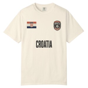 May include: Cream-colored t-shirt featuring the word "CROATIA" in black block letters. The shirt has a Croatian flag patch on the left chest and a soccer-themed crest on the right chest.