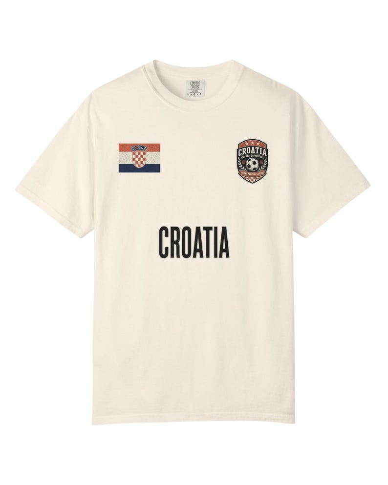 May include: Cream-colored t-shirt featuring the word "CROATIA" in black block letters. The shirt has a Croatian flag patch on the left chest and a soccer-themed crest on the right chest.