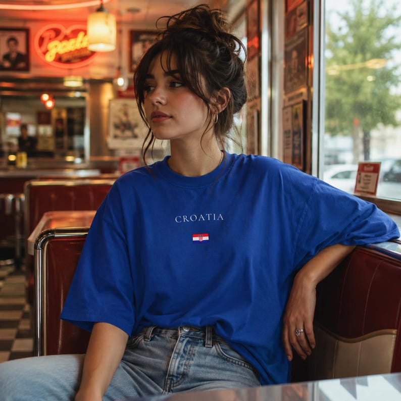 Peut inclure: Un t-shirt bleu avec le mot "CROATIA" et un petit drapeau croate. Le t-shirt est de couleur bleu uni et a un col rond. Le t-shirt est port&eacute; avec un jean bleu.