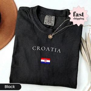 Peut inclure: T-shirt noir pli&eacute; avec le mot "CROATIA" en blanc et un petit drapeau croate brod&eacute;. Le col affiche une &eacute;tiquette Comfort Colors. Un collier pendentif dor&eacute; est pos&eacute; sur le tissu.