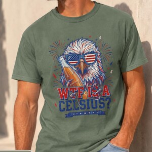 May include: A moss green short-sleeve t-shirt featuring a graphic of a bald eagle wearing American flag sunglasses and holding a beer bottle. The text reads "WTF IS A CELSIUS?" in red and blue lettering, surrounded by firework illustrations.