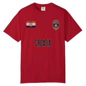May include: Red t-shirt featuring the word "CROATIA" in black lettering. The shirt has a Croatian flag patch on the left chest and a soccer-themed emblem on the right chest. The shirt is a short-sleeved crew neck.