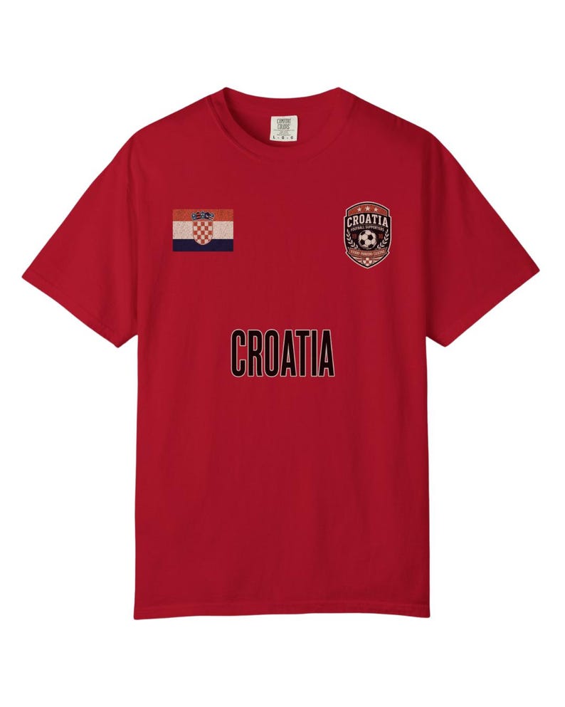 May include: Red t-shirt featuring the word "CROATIA" in black lettering. The shirt has a Croatian flag patch on the left chest and a soccer-themed emblem on the right chest. The shirt is a short-sleeved crew neck.