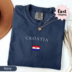 Peut inclure: T-shirt bleu marine avec le mot "CROATIA" en blanc et un petit drapeau croate. Le t-shirt a un col rond et une &eacute;tiquette "COMFORT COLORS". Un collier en or avec un pendentif est pos&eacute; sur le t-shirt.