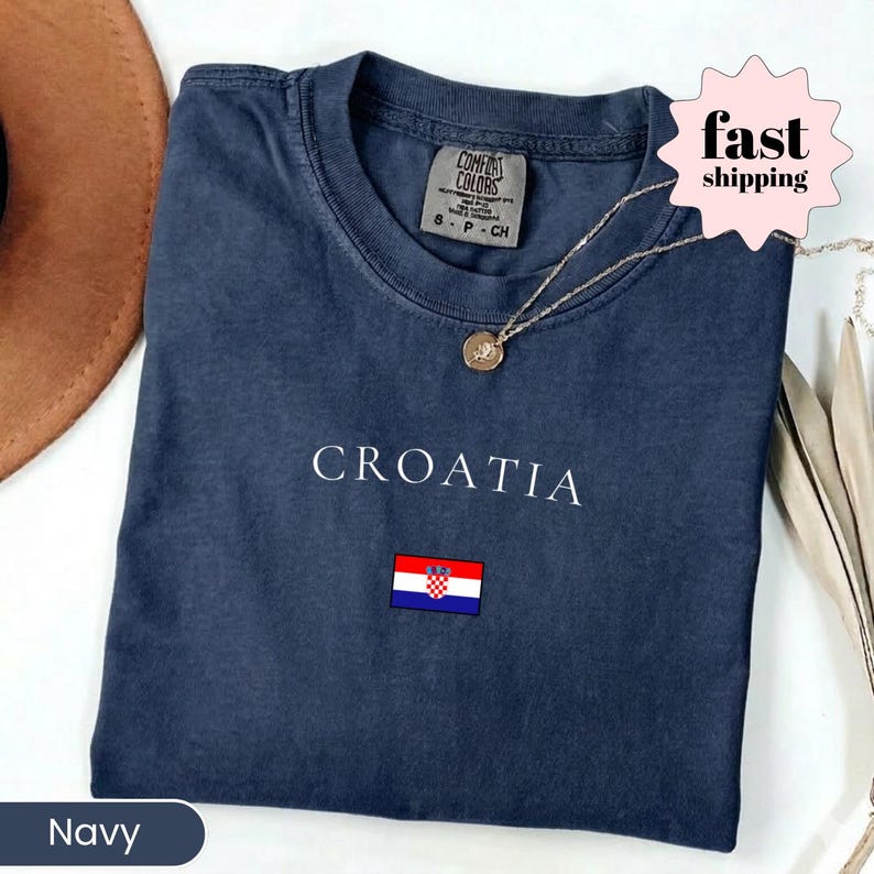Peut inclure: T-shirt bleu marine avec le mot "CROATIA" en blanc et un petit drapeau croate. Le t-shirt a un col rond et une &eacute;tiquette "COMFORT COLORS". Un collier en or avec un pendentif est pos&eacute; sur le t-shirt.