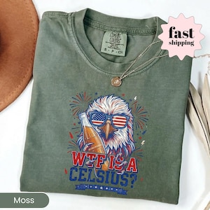 May include: A moss green crew neck t-shirt featuring a graphic of a bald eagle wearing American flag sunglasses and holding a glass bottle. The text reads "WTF IS A CELSIUS?" in blue and red lettering, surrounded by red and blue firework illustrations.