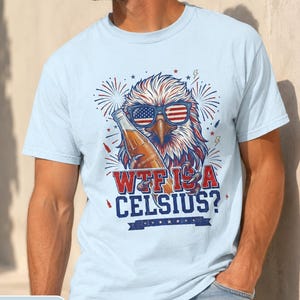 May include: A light blue t-shirt featuring a graphic of a bald eagle wearing American flag sunglasses and holding a glass bottle. The text reads WTF IS A CELSIUS? in red and blue lettering, surrounded by firework illustrations and small lightning bolts.
