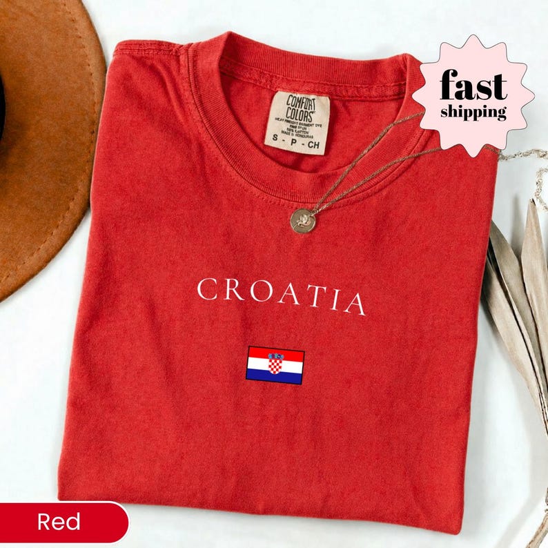 Peut inclure: Un t-shirt rouge avec le mot "CROATIA" en blanc et un petit drapeau croate. Le t-shirt a un col rond et des manches courtes. Un collier dor&eacute; avec un pendentif est drap&eacute; sur le t-shirt.