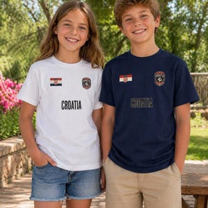 May include: Two children wearing Croatia soccer t-shirts. One shirt is white, the other is navy blue. Both shirts have the Croatian flag and a soccer emblem. The word "CROATIA" is printed in black below the emblem.