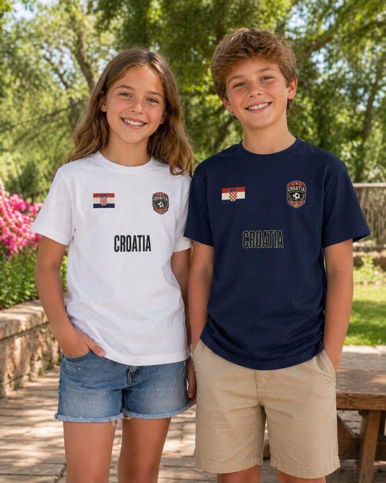 May include: Two children wearing Croatia soccer t-shirts. One shirt is white, the other is navy blue. Both shirts have the Croatian flag and a soccer emblem. The word "CROATIA" is printed in black below the emblem.