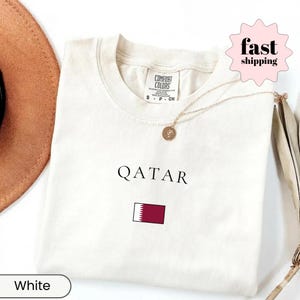 Może przedstawiać: Biała koszulka z czarnym napisem "QATAR" i flagą Kataru. Koszulka ma okrągły dekolt i kr&oacute;tkie rękawy. Złoty naszyjnik z wisiorkiem jest ułożony na koszulce.