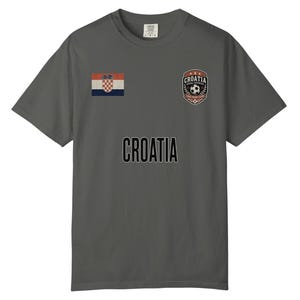 May include: A gray t-shirt featuring the word "CROATIA" in large, black letters. The shirt has a small Croatian flag on the left chest and a soccer-themed emblem on the right chest. The shirt is a comfortable, casual style.