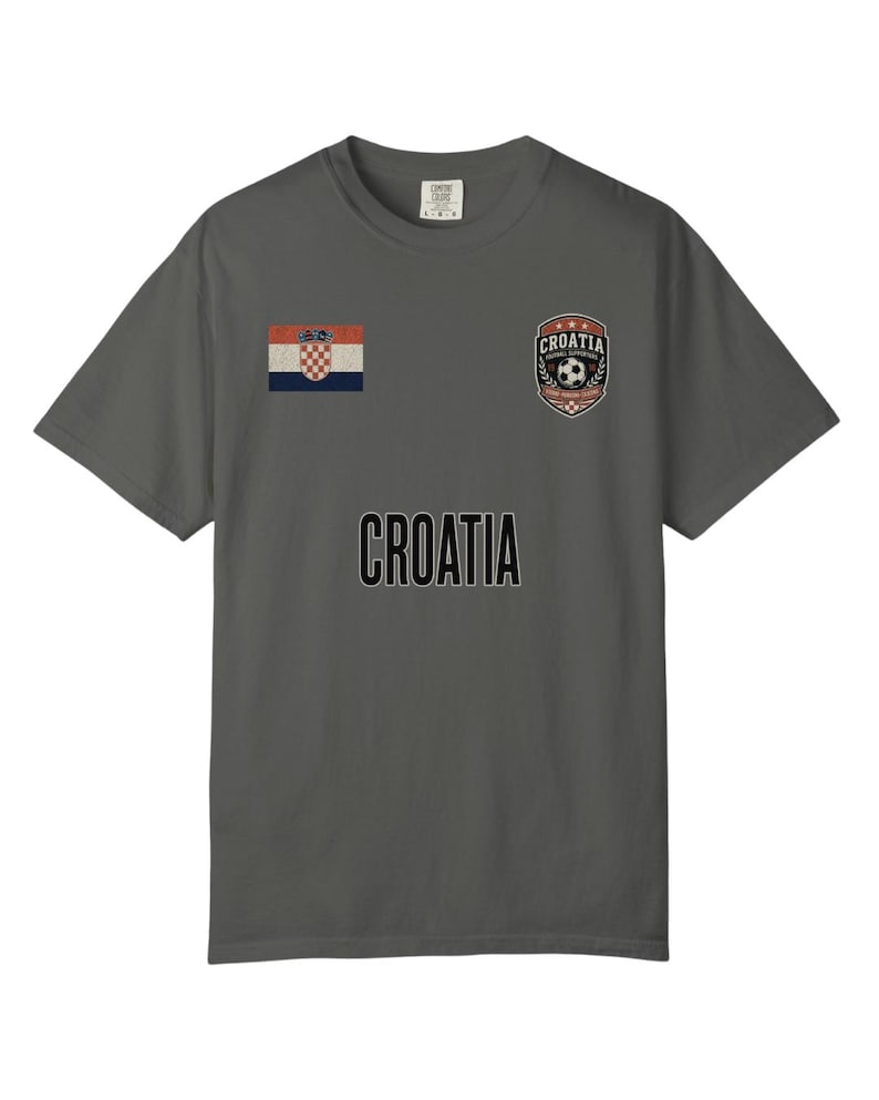 May include: A gray t-shirt featuring the word "CROATIA" in large, black letters. The shirt has a small Croatian flag on the left chest and a soccer-themed emblem on the right chest. The shirt is a comfortable, casual style.