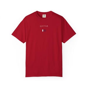 Może przedstawiać: Czerwony t-shirt z białym napisem "QATAR" nad małą grafiką flagi. Słowo "RED" jest wydrukowane pogrubioną czcionką na dole. Koszulka wykonana jest z miękkiego materiału.