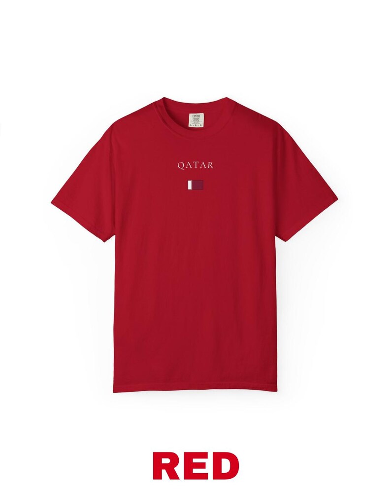 Może przedstawiać: Czerwony t-shirt z białym napisem "QATAR" nad małą grafiką flagi. Słowo "RED" jest wydrukowane pogrubioną czcionką na dole. Koszulka wykonana jest z miękkiego materiału.