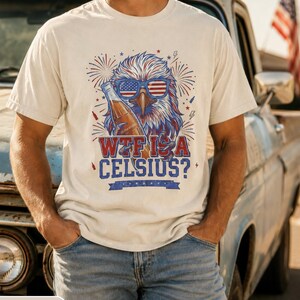May include: An ivory short-sleeve t-shirt featuring a graphic of a bald eagle wearing American flag sunglasses and holding a glass bottle. The design includes red and blue fireworks, lightning bolts, and the text "WTF IS A CELSIUS?" in bold.