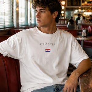 Peut inclure: T-shirt blanc avec le mot "CROATIA" imprim&eacute; au-dessus d'un petit drapeau croate. Le t-shirt a des manches courtes et est fait d'une mati&egrave;re douce. Le t-shirt est port&eacute; par une personne.