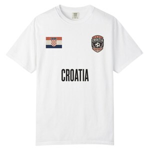 May include: White t-shirt featuring the word "CROATIA" in large black letters. The shirt has a Croatian flag patch on the left chest and a soccer-themed emblem on the right chest. The shirt is a casual wear item.